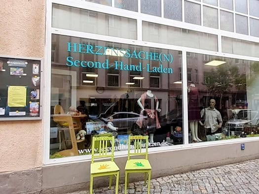 Schaufenster des Second-Hand-Ladens Herzenssache(n) in Bautzen mit Schaufenster, Kleidung, Spielzeug und bunten Stühlen vor dem Eingang.