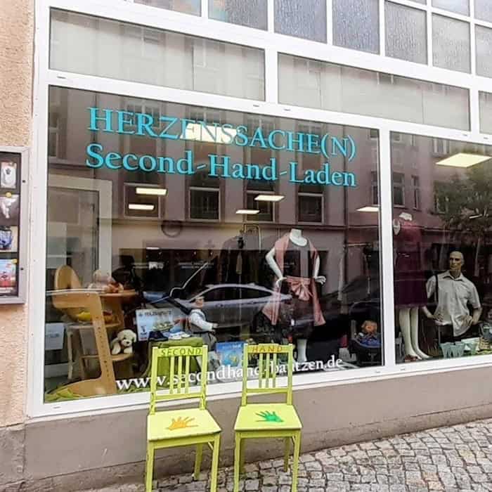 Schaufenster des Second-Hand-Ladens Herzenssache(n) in Bautzen mit Schaufenster, Kleidung, Spielzeug und bunten Stühlen vor dem Eingang.