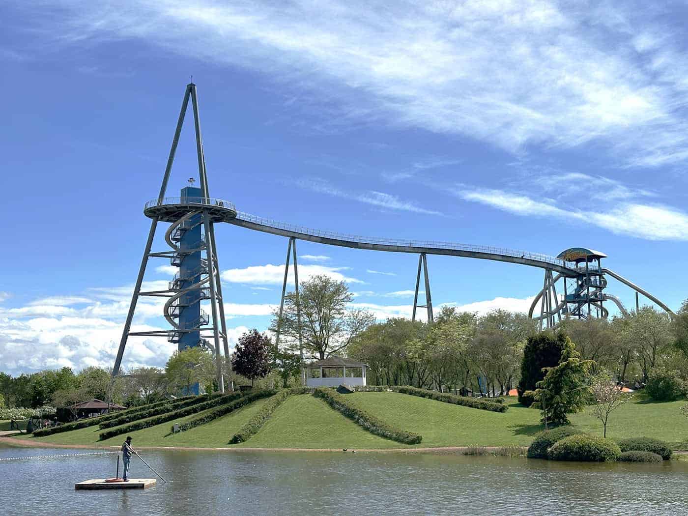 Ansicht Skywalk und Pyramide im Sonnenlandpark Lichtenau. Weitwinkelaufnahme des Sonnenlandparks Lichtenau mit der neuen, weitläufigen Skywalk-Konstruktion. Ein hoher, moderner pyramidenförmiger Turm aus Metall mit außenliegender Wendeltreppe (rechts) ist über eine lange, leicht ansteigende stählerne Brücke mit einem zweiten Turm verbunden (links). Darunter befinden sich Bäume, eine grüne Rasenfläche und ein großer, dunkelbrauner Teich mit einer Person auf einem schwimmenden Floß unter einem strahlend blauen Himmel mit leichten Wolken.