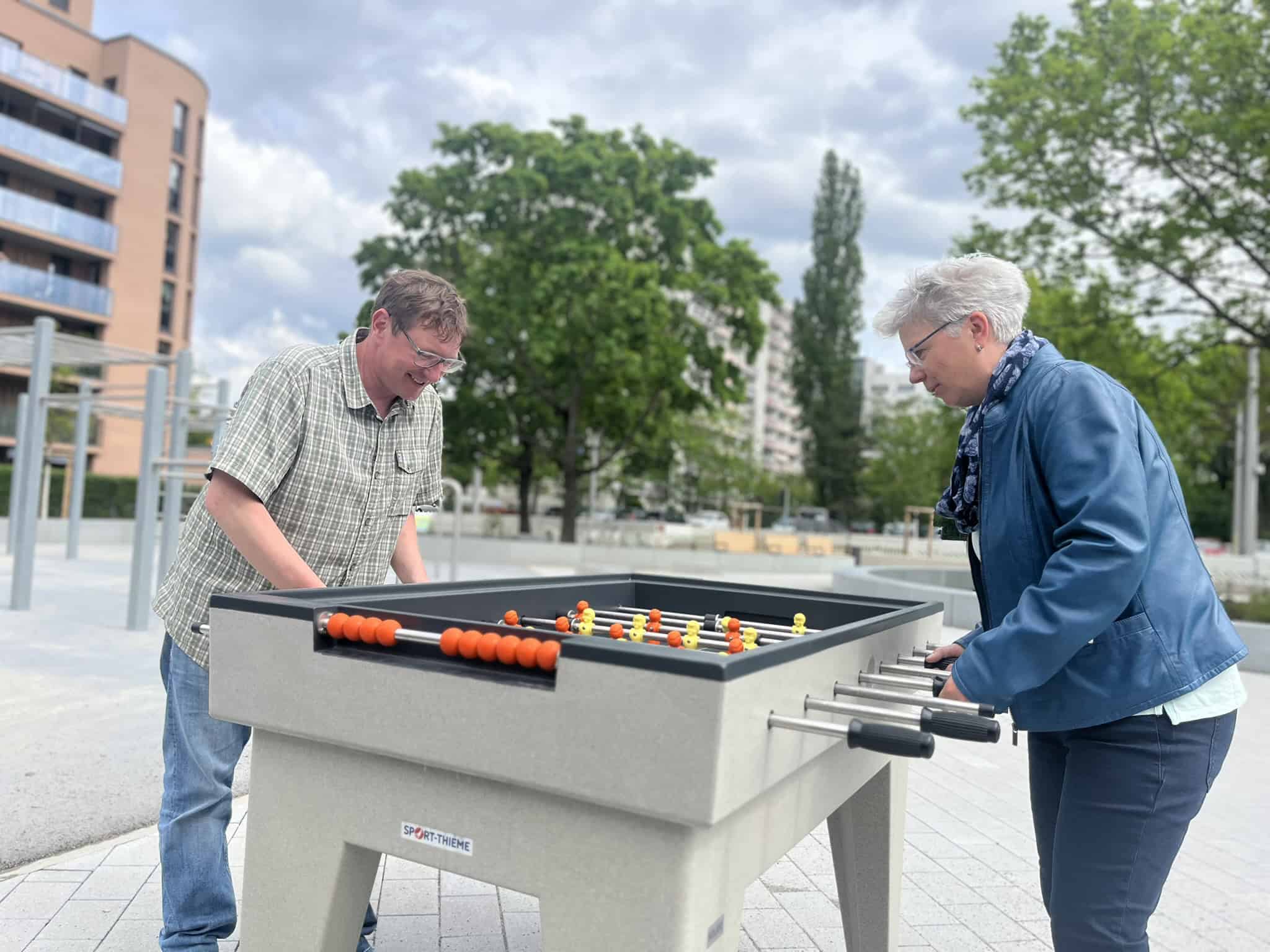 Eine Frau in blauer Lederjacke und ein Mann in einem grün-karierten Hemd spielen an einem massiven Outdoor-Tischkicker aus grauem Beton.