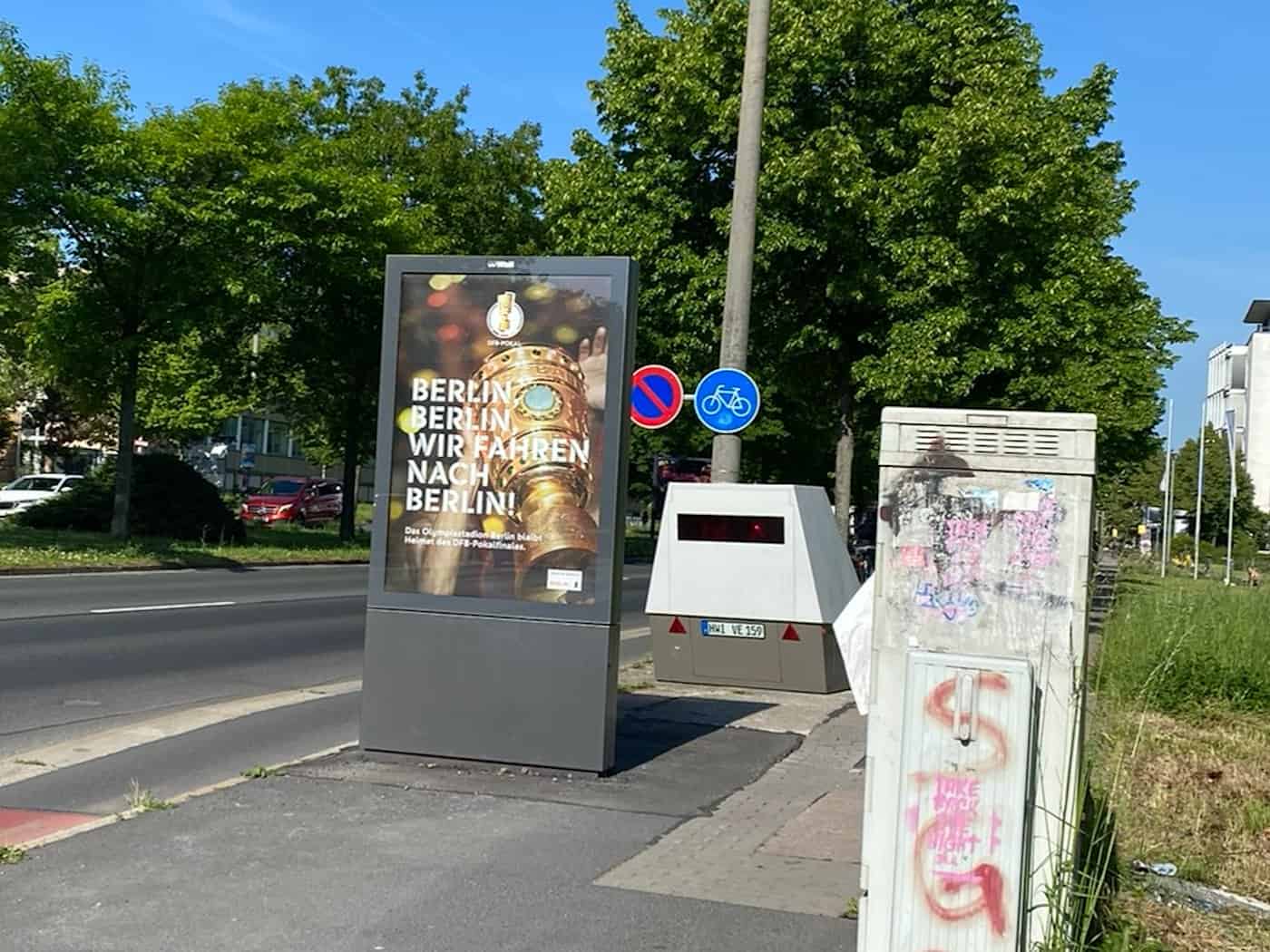 Ein grauer Blitzer-Messanhänger steht strategisch versteckt direkt hinter einer großen Werbetafel auf dem Gehweg am Zelleschen Weg in Dresden. Die Werbetafel zeigt ein Plakat zum DFB-Pokalfinale mit der Aufschrift „Berlin, Berlin, wir fahren nach Berlin!“. Im Vordergrund ist ein grauer, mit Graffiti
