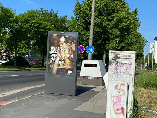 Ein grauer Blitzer-Messanhänger steht strategisch versteckt direkt hinter einer großen Werbetafel auf dem Gehweg am Zelleschen Weg in Dresden. Die Werbetafel zeigt ein Plakat zum DFB-Pokalfinale mit der Aufschrift „Berlin, Berlin, wir fahren nach Berlin!“. Im Vordergrund ist ein grauer, mit Graffiti
