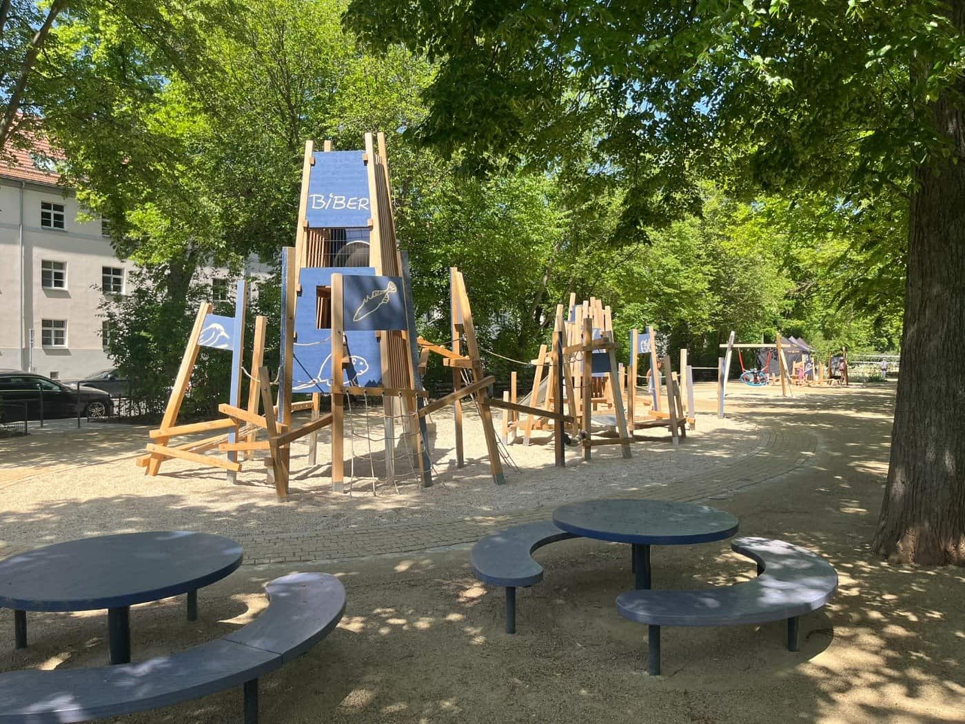 Zwei runde Picknicktische mit integrierten Bänken auf hellem Kiesboden im Toeplerpark Tolkewitz, im Hintergrund ist ein Spielgerät für kleinere Kinder zu sehen.