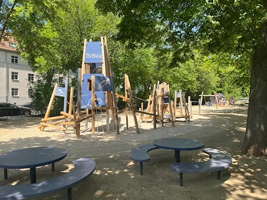Zwei runde Picknicktische mit integrierten Bänken auf hellem Kiesboden im Toeplerpark Tolkewitz, im Hintergrund ist ein Spielgerät für kleinere Kinder zu sehen.