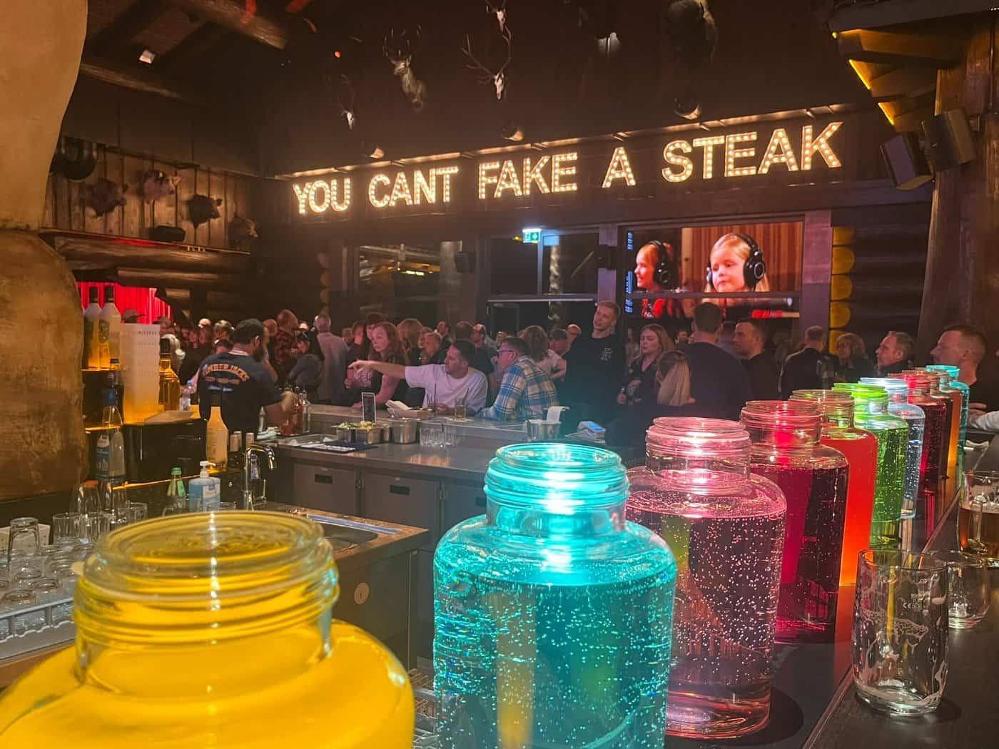 Innenansicht einer belebten Bar im Timberjacks Bannewitz. Im Vordergrund stehen bunte, leuchtende Glasbehälter mit Flüssigkeit. Über den Köpfen der Gäste leuchtet der Schriftzug „YOU CAN’T FAKE A STEAK“.
