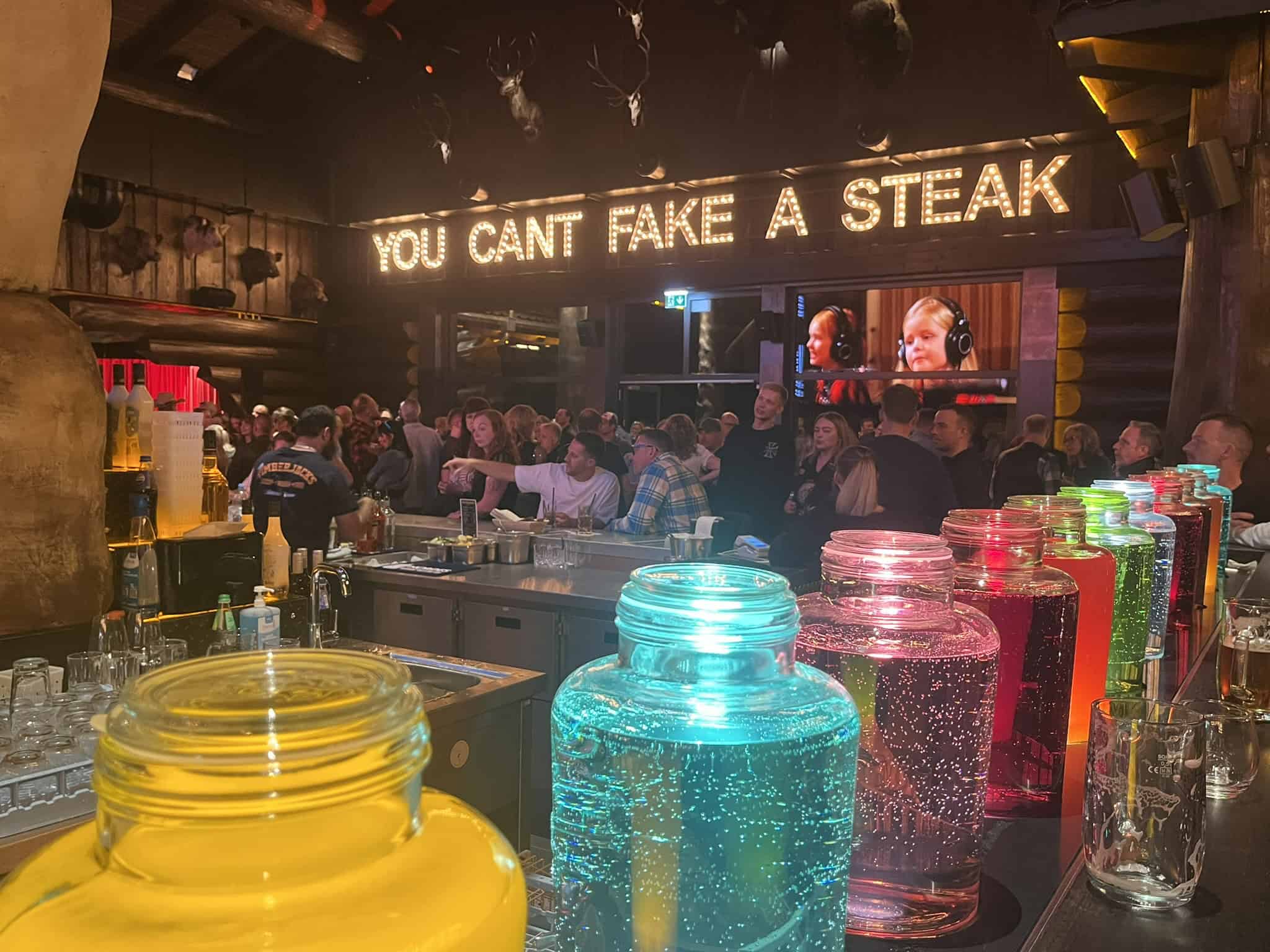 Innenansicht einer belebten Bar im Timberjacks Bannewitz. Im Vordergrund stehen bunte, leuchtende Glasbehälter mit Flüssigkeit. Über den Köpfen der Gäste leuchtet der Schriftzug „YOU CAN’T FAKE A STEAK“.
