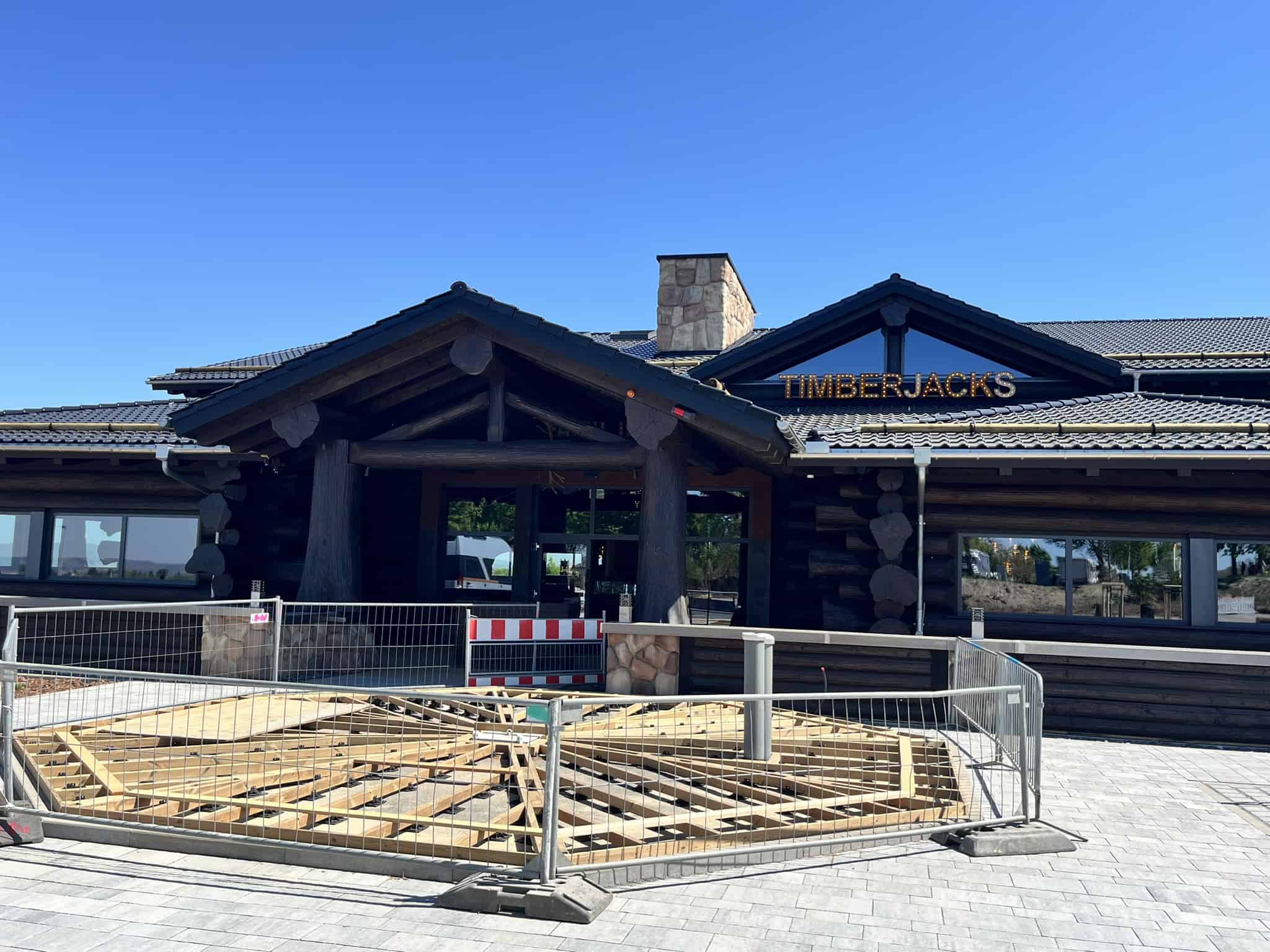 Außenansicht des Timberjacks Restaurants in Bannewitz (B170). Ein dunkles Blockhaus mit dem beleuchteten Schriftzug "TIMBERJACKS" am Giebel. Im Vordergrund sind Bauzäune und eine hölzerne Unterkonstruktion für eine Terrasse zu sehen. Blauer, wolkenloser Himmel.