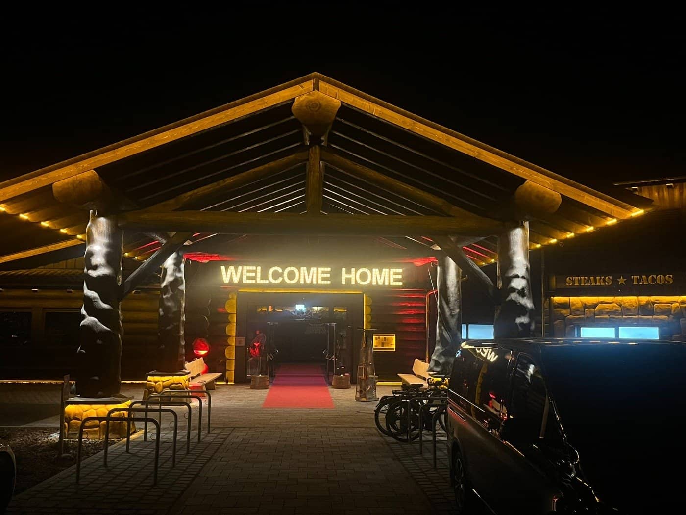 Nachtaufnahme des Eingangs eines großen Blockhauses. Ein hölzernes Vordach wird von massiven Baumstämmen getragen. Über dem Eingang leuchtet der Schriftzug „WELCOME HOME“ in warmem Licht. Ein roter Teppich führt ins Innere.