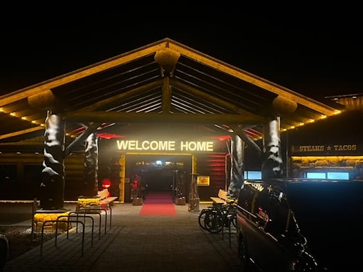 Nachtaufnahme des Eingangs eines großen Blockhauses. Ein hölzernes Vordach wird von massiven Baumstämmen getragen. Über dem Eingang leuchtet der Schriftzug „WELCOME HOME“ in warmem Licht. Ein roter Teppich führt ins Innere.