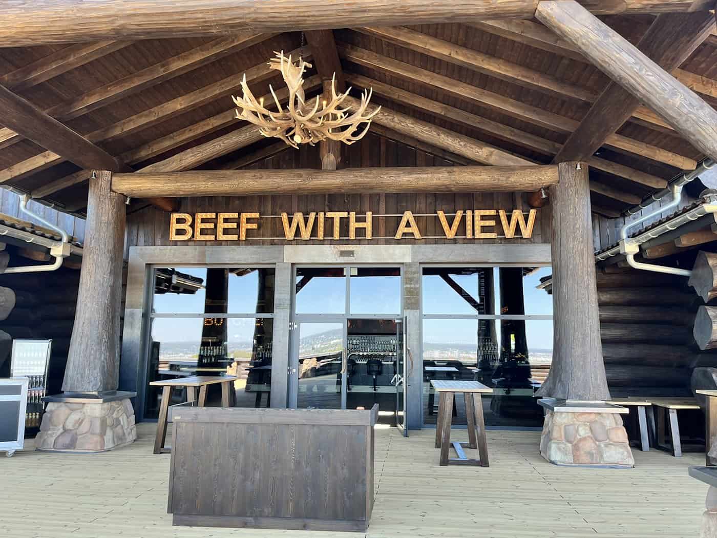 Außenansicht der Terrasse eines rustikalen Holzgebäudes. Über den Glastüren hängt ein leuchtender Schriftzug „BEEF WITH A VIEW“. Von der Decke hängt ein großer Kronleuchter aus Geweihen.