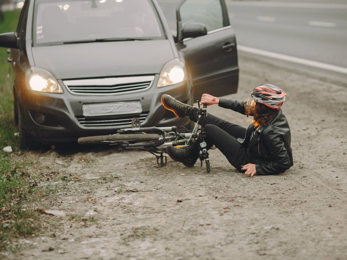 Symbolbild eines Verkehrsunfalls: Eine Person mit Fahrradhelm sitzt nach einem Sturz neben ihrem Fahrrad auf dem Boden vor einem Auto mit eingeschalteten Scheinwerfern.