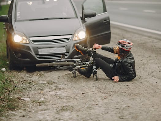 Symbolbild eines Verkehrsunfalls: Eine Person mit Fahrradhelm sitzt nach einem Sturz neben ihrem Fahrrad auf dem Boden vor einem Auto mit eingeschalteten Scheinwerfern.