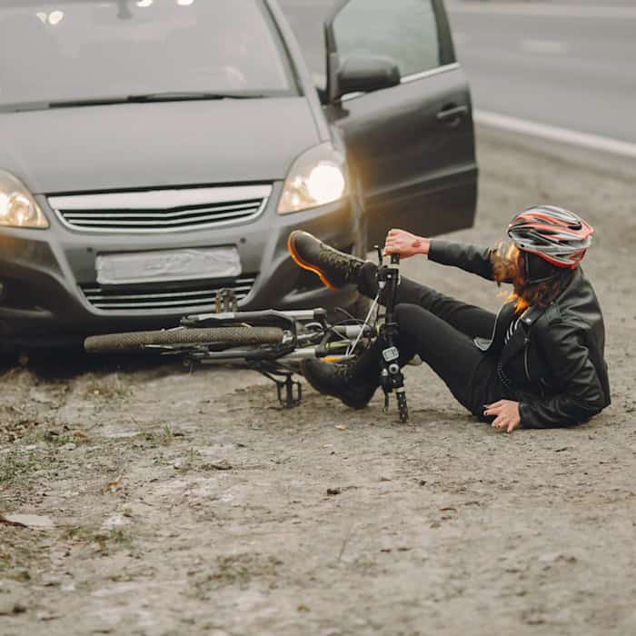 Symbolbild eines Verkehrsunfalls: Eine Person mit Fahrradhelm sitzt nach einem Sturz neben ihrem Fahrrad auf dem Boden vor einem Auto mit eingeschalteten Scheinwerfern.