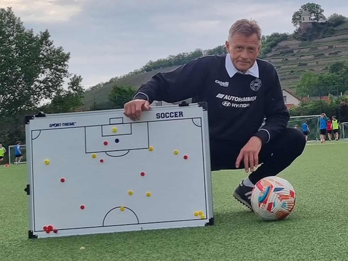 Foto: Ein Mann hockt auf Kunstrasen, vor sich ein Fußball und ein Whiteboard mit der Darstellung eines Spielfelds