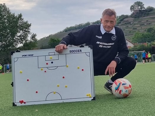 Foto: Ein Mann hockt auf Kunstrasen, vor sich ein Fußball und ein Whiteboard mit der Darstellung eines Spielfelds