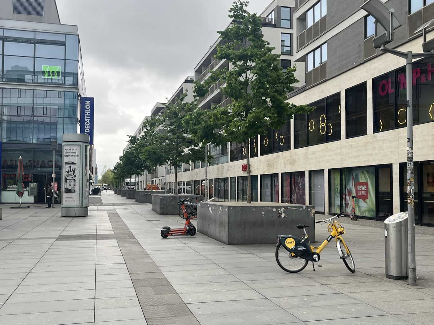 Eine Weitwinkelaufnahme auf Straßenebene vom gepflasterten Wiener Platz in Dresden mit Blick nach Süden zum Hauptbahnhof unter einem bewölkten Himmel. Große Betonkübel mit Bäumen säumen den Fußgängerbereich auf beiden Seiten. Im Vordergrund stehen ein gelbes Leihfahrrad und ein orangefarbener E-Scooter. Moderne Geschäftsgebäude mit Logos von Decathlon und Rewe rahmen den Platz ein. Das Bild zeigt die Elemente, die bis zum Sommer 2026 entfernt werden sollen.