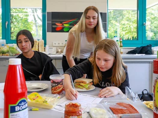 Eine Studentin und zwei Schülerinnen arbeiten in der Schulküche der Richard-von-Schlieben-Oberschule in Zittau an einem Projekt zum Thema Ernährung. Auf dem Tisch stehen Zutaten für ein Sandwich und einfertiges Essen. Eine Schülerin bearbeitet ein Aufgabe