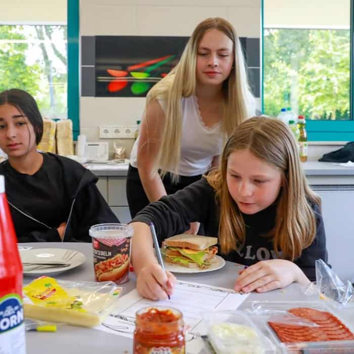 Eine Studentin und zwei Schülerinnen arbeiten in der Schulküche der Richard-von-Schlieben-Oberschule in Zittau an einem Projekt zum Thema Ernährung. Auf dem Tisch stehen Zutaten für ein Sandwich und einfertiges Essen. Eine Schülerin bearbeitet ein Aufgabe