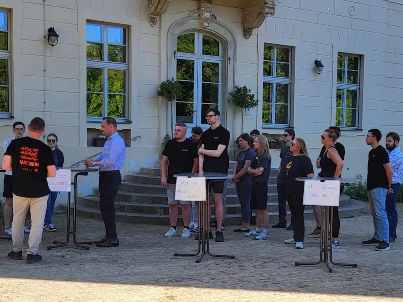 Etwa ein Dutzend jüngere Menschen stehen hinter Stehtischen; sie schauen auf mehrere Personen am linken Bildrand, die sich unterhalten.
