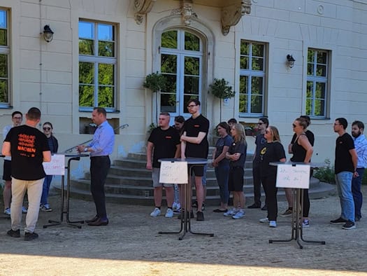 Etwa ein Dutzend jüngere Menschen stehen hinter Stehtischen; sie schauen auf mehrere Personen am linken Bildrand, die sich unterhalten.