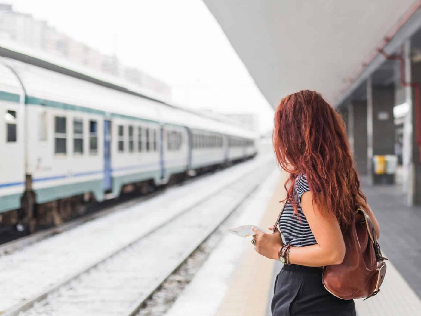 Frau mit Rucksack und Smartphone steht auf einem Bahnsteig, im Hintergrund fährt ein Zug ein.