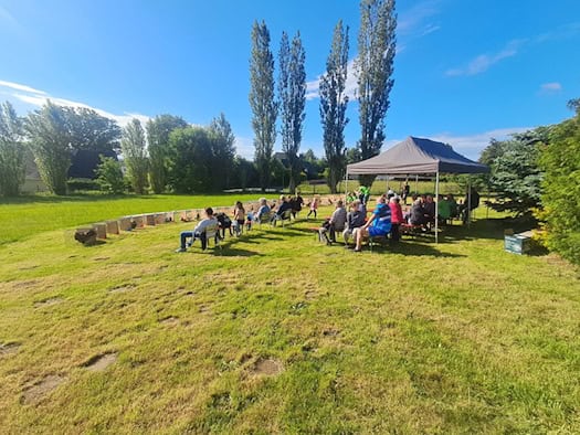 Foto: Auf einer Wiese bei blauen Himmel und umsäumt von grünen Bäumen sitzen auf Bierbänken und Stühlen aufgereiht knapp zwei Dutzend Menschen. Vor ihnen in einer Reihe stehen Käfige mit Hähnen darin.