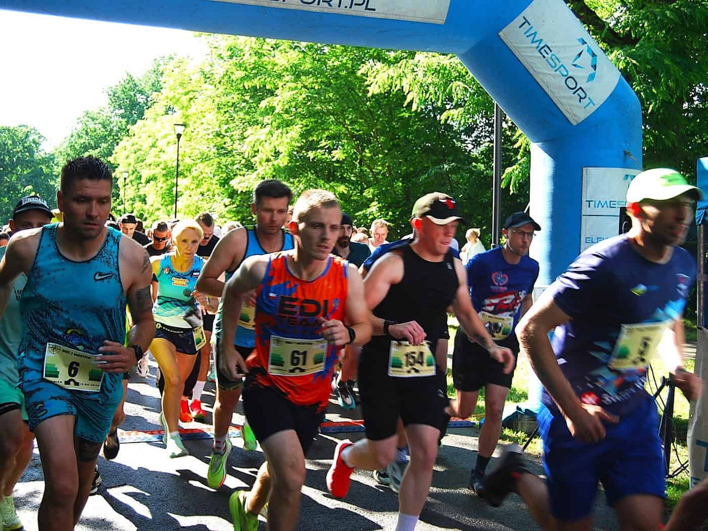 Foto: Dutzende Läuferinnen und Läufer in verschiedenfarbigen kurzen Sportsachen mit Nummern auf dem Bau passieren rennend ein mit Luft aufgeblasenes Tor mit Werbebannern. Im Hintergrund früne Bäume von der Sonne beschienen.