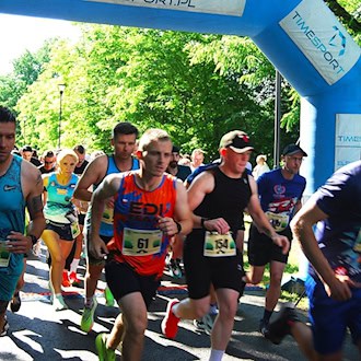 Foto: Dutzende Läuferinnen und Läufer in verschiedenfarbigen kurzen Sportsachen mit Nummern auf dem Bau passieren rennend ein mit Luft aufgeblasenes Tor mit Werbebannern. Im Hintergrund früne Bäume von der Sonne beschienen.