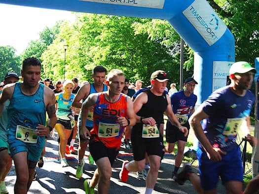 Foto: Dutzende Läuferinnen und Läufer in verschiedenfarbigen kurzen Sportsachen mit Nummern auf dem Bau passieren rennend ein mit Luft aufgeblasenes Tor mit Werbebannern. Im Hintergrund früne Bäume von der Sonne beschienen.