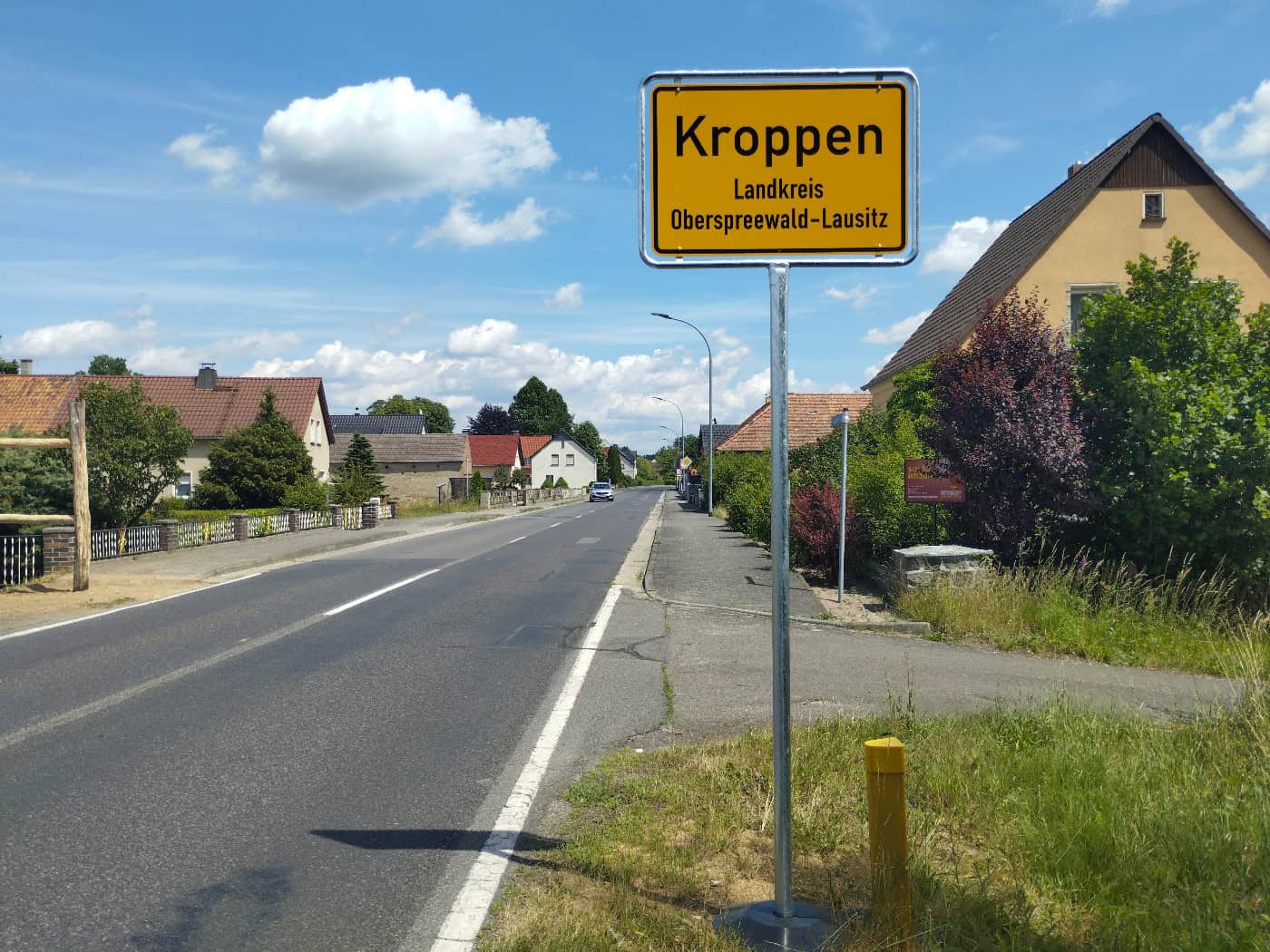 Foto: Ortseingangsschild Kroppen an einer Straße, die an dörflichen Häusern und Bäumen vorbeiführt. Der Himmel ist heiter.