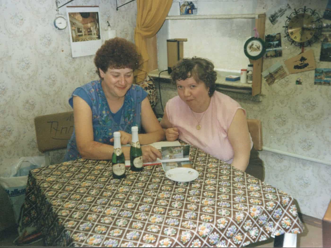 Foto: Zwei Frauen mit kurzem Haar sitzen lächelnd an einem kleinen Tisch mit gemusterter Tischdecke, vor Ihnen ein Fotoalbum und zwei kleine Flaschen Sekt. Es hängen Bilder, Kalender und eine Uhr an der Wand, die gemustert tapeziert ist.