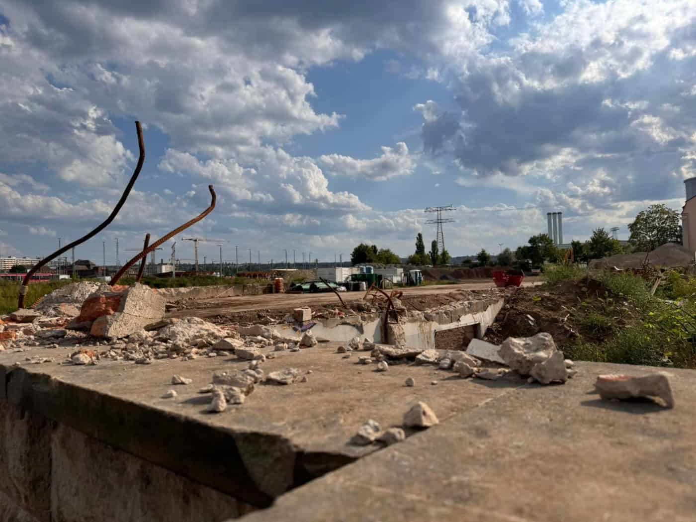 Blick über eine staubige Baustelle mit Betonbrocken und verbogenem, rostigen Bewehrungsstahl im Vordergrund. Im Hintergrund sieht man ein flaches, freies Areal, Container, Strommasten und Fabrikschornsteine unter bewölktem Himmel.