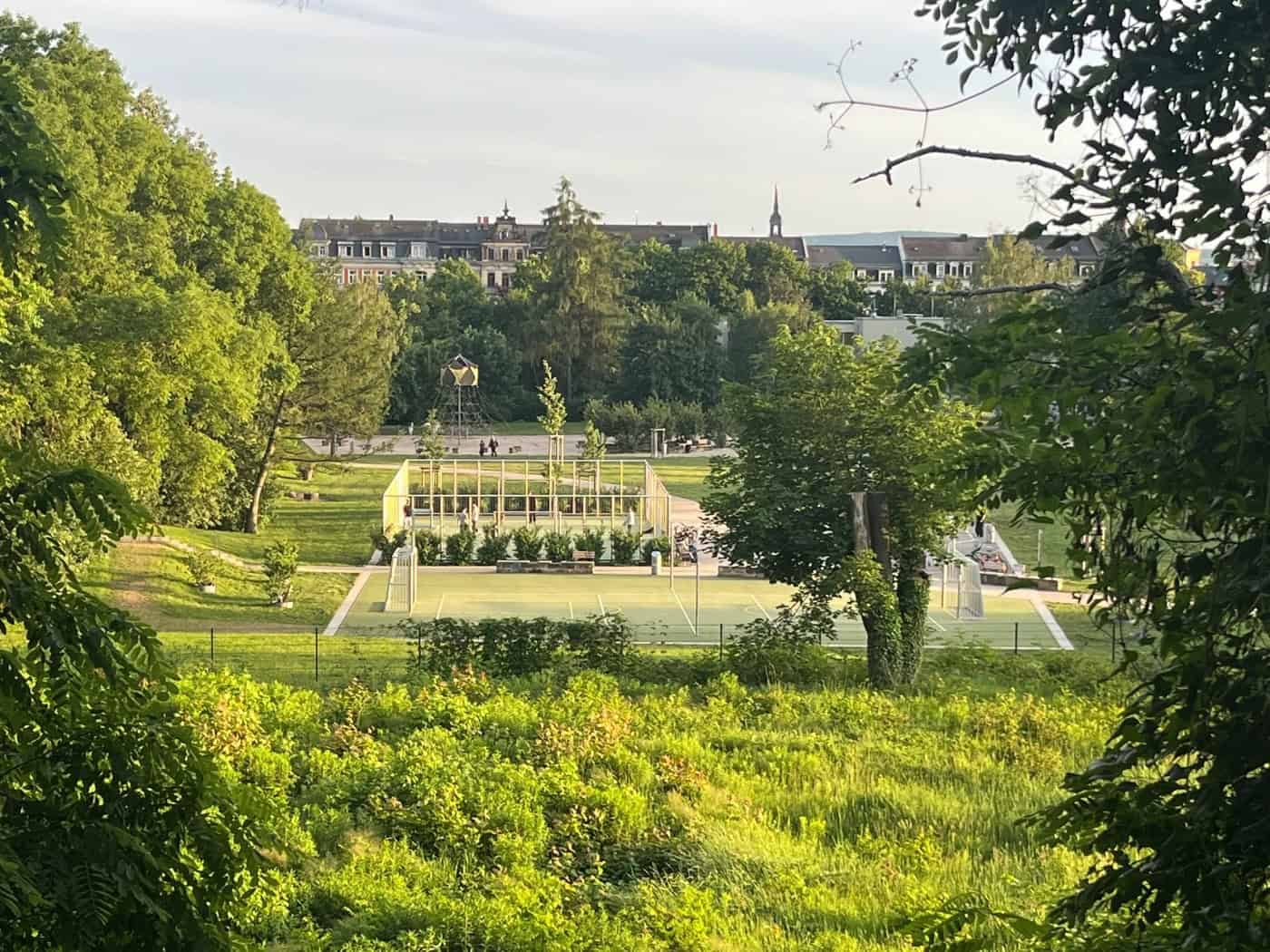 Blick von einem erhöhten, von grünen Sträuchern gesäumten Standpunkt auf die modernen Sport- und Freizeitanlagen in der Westerweiterung des Alaunparks. Im Hintergrund erstrecken sich grüne Bäume und die Häuserfassaden der Dresdner Neustadt unter einem hellen Himmel.