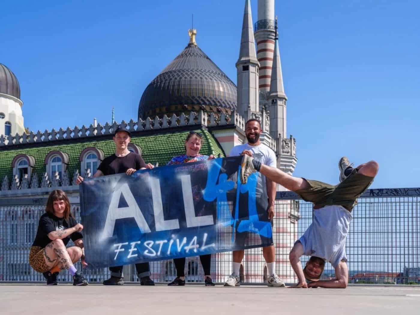 Ein Gruppenfoto von fünf Personen in Freizeitkleidung und Streetwear, die auf einem Parkdeck bei Tageslicht posieren. Direkt hinter ihnen ragt das markante Gebäude der Yenidze mit seiner gläsernen Kuppel empor. Der Himmel ist blau mit leichten Wolken.