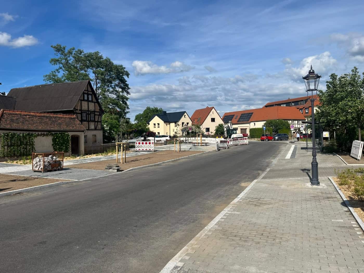 Blick auf die neu asphaltierte Straße und die gepflasterten Gehwege des historischen Dorfangers Altdobritz in Dresden. Auf der linken Seite steht ein gut erhaltenes, altes Fachwerkhaus, auf der rechten Seite eine klassische, schwarze Kandelaber-Laterne. Im Hintergrund sind vereinzelte Baustellenabsp