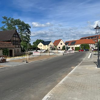 Blick auf die neu asphaltierte Straße und die gepflasterten Gehwege des historischen Dorfangers Altdobritz in Dresden. Auf der linken Seite steht ein gut erhaltenes, altes Fachwerkhaus, auf der rechten Seite eine klassische, schwarze Kandelaber-Laterne. Im Hintergrund sind vereinzelte Baustellenabsp