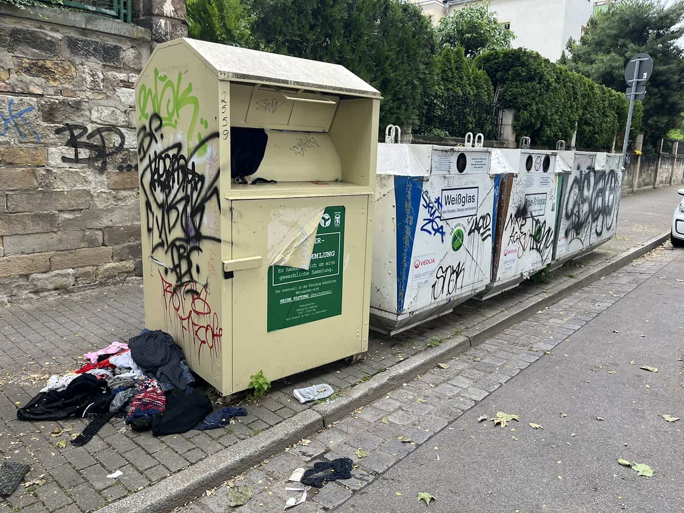 Ein beigefarbener, mit Graffiti bespühter Altkleidercontainer am Carusufer in Dresden-Neustadt, neben dem Kleidung auf dem Gehweg liegt.