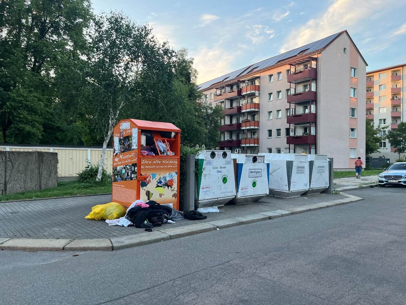 Ein vollgestopfter oranger Altkleidercontainer an der Falkenstraße in Dresden mit davor aufgetürmten Kleiderhaufen vor einem mehrstöckigen Wohnblock.