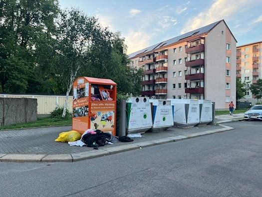 Ein vollgestopfter oranger Altkleidercontainer an der Falkenstraße in Dresden mit davor aufgetürmten Kleiderhaufen vor einem mehrstöckigen Wohnblock.