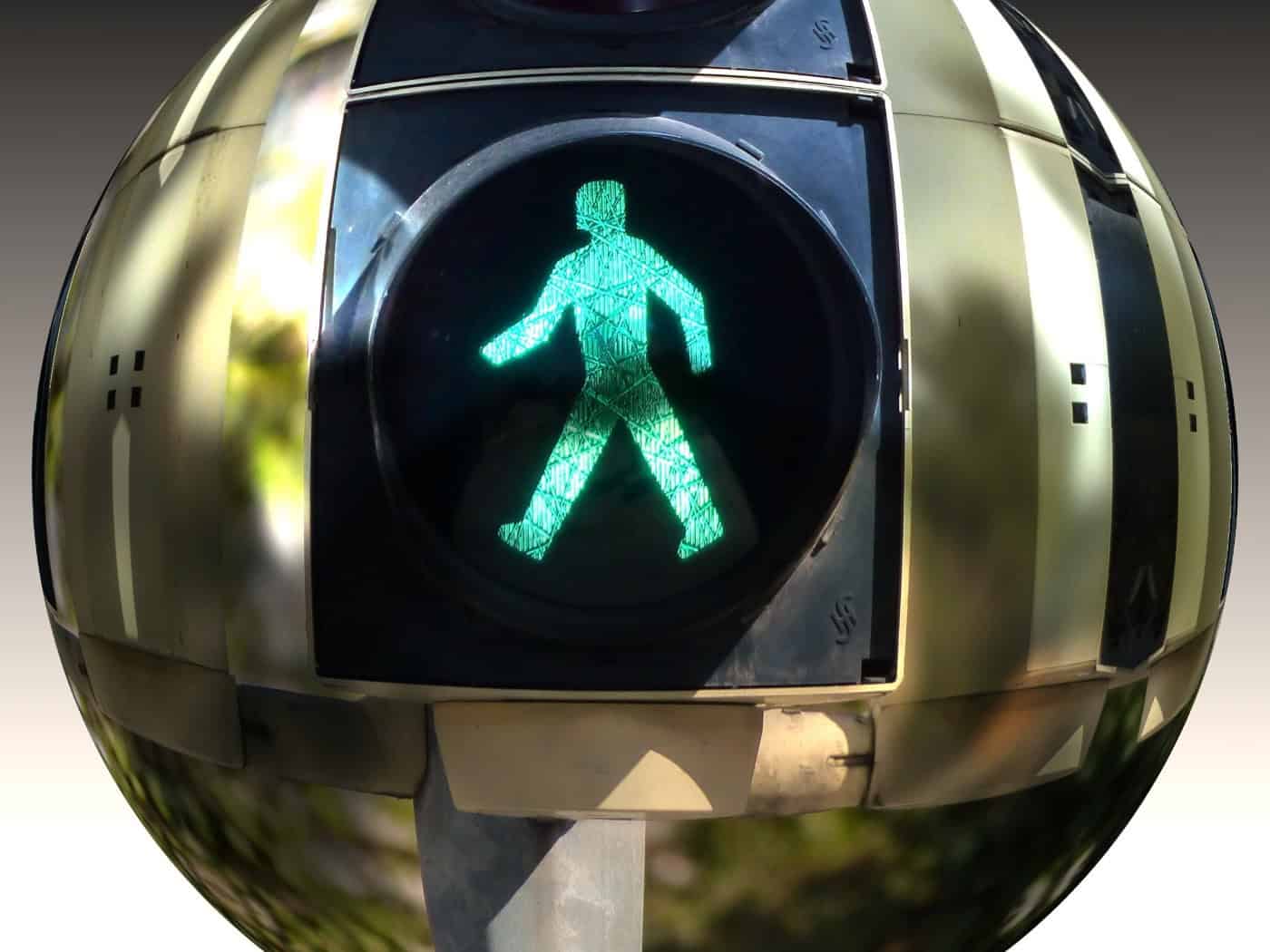 Eine Nahaufnahme einer grünen Fußgängerampel, die in einer kreisrunden, spiegelnden Glaskugel stark verzerrt dargestellt wird. Das leuchtende grüne Ampelmännchen befindet sich exakt im Zentrum der Kugel.