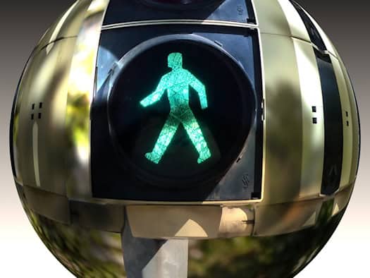 Eine Nahaufnahme einer grünen Fußgängerampel, die in einer kreisrunden, spiegelnden Glaskugel stark verzerrt dargestellt wird. Das leuchtende grüne Ampelmännchen befindet sich exakt im Zentrum der Kugel.