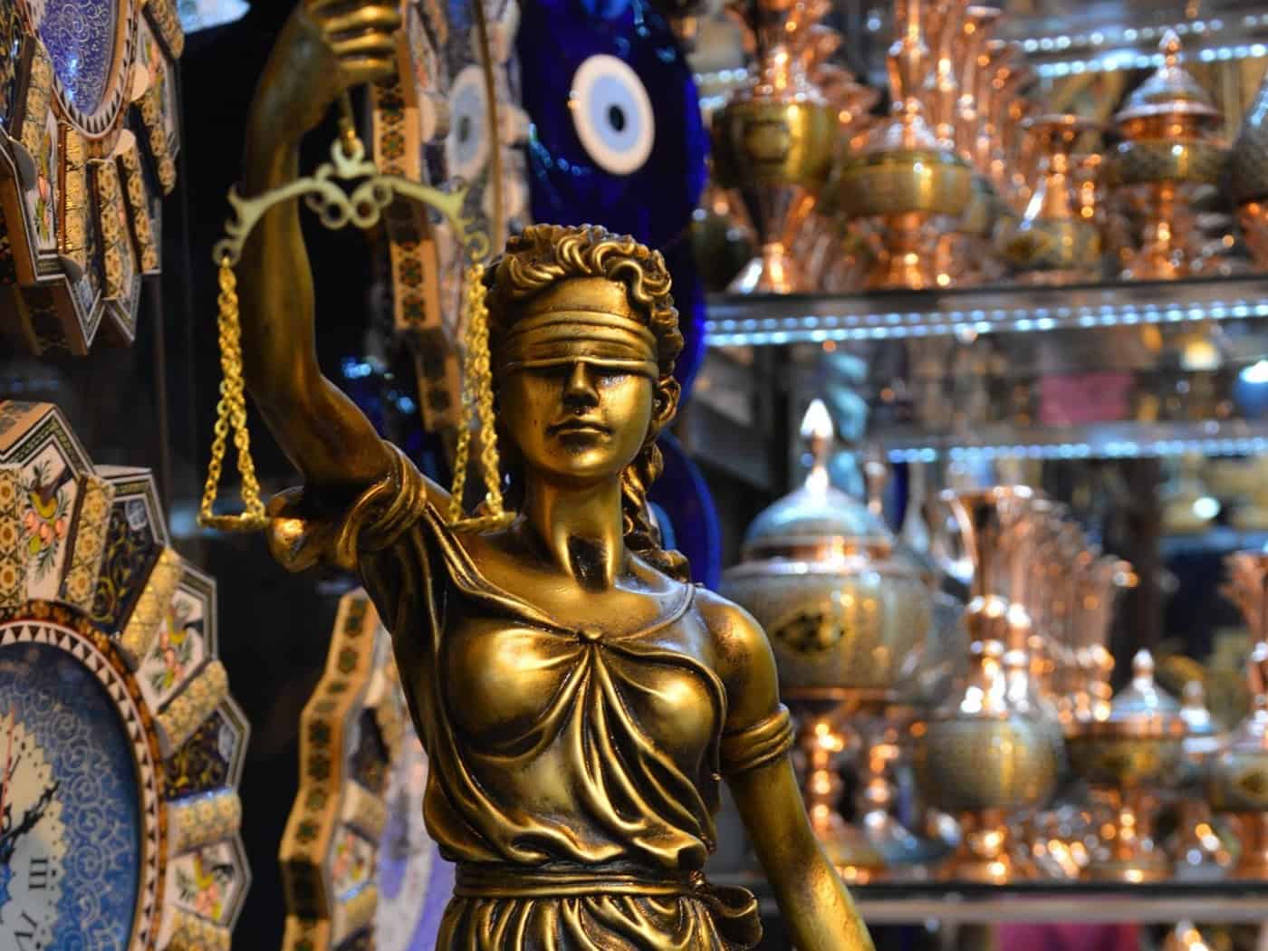 Eine goldene Justitia-Statue mit Augenbinde, Waage und Schwert steht als Symbol für das Rechtssystem im Vordergrund. Im unscharfen Hintergrund befinden sich dekorative orientalische Gefäße und Uhren.