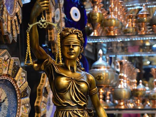 Eine goldene Justitia-Statue mit Augenbinde, Waage und Schwert steht als Symbol für das Rechtssystem im Vordergrund. Im unscharfen Hintergrund befinden sich dekorative orientalische Gefäße und Uhren.