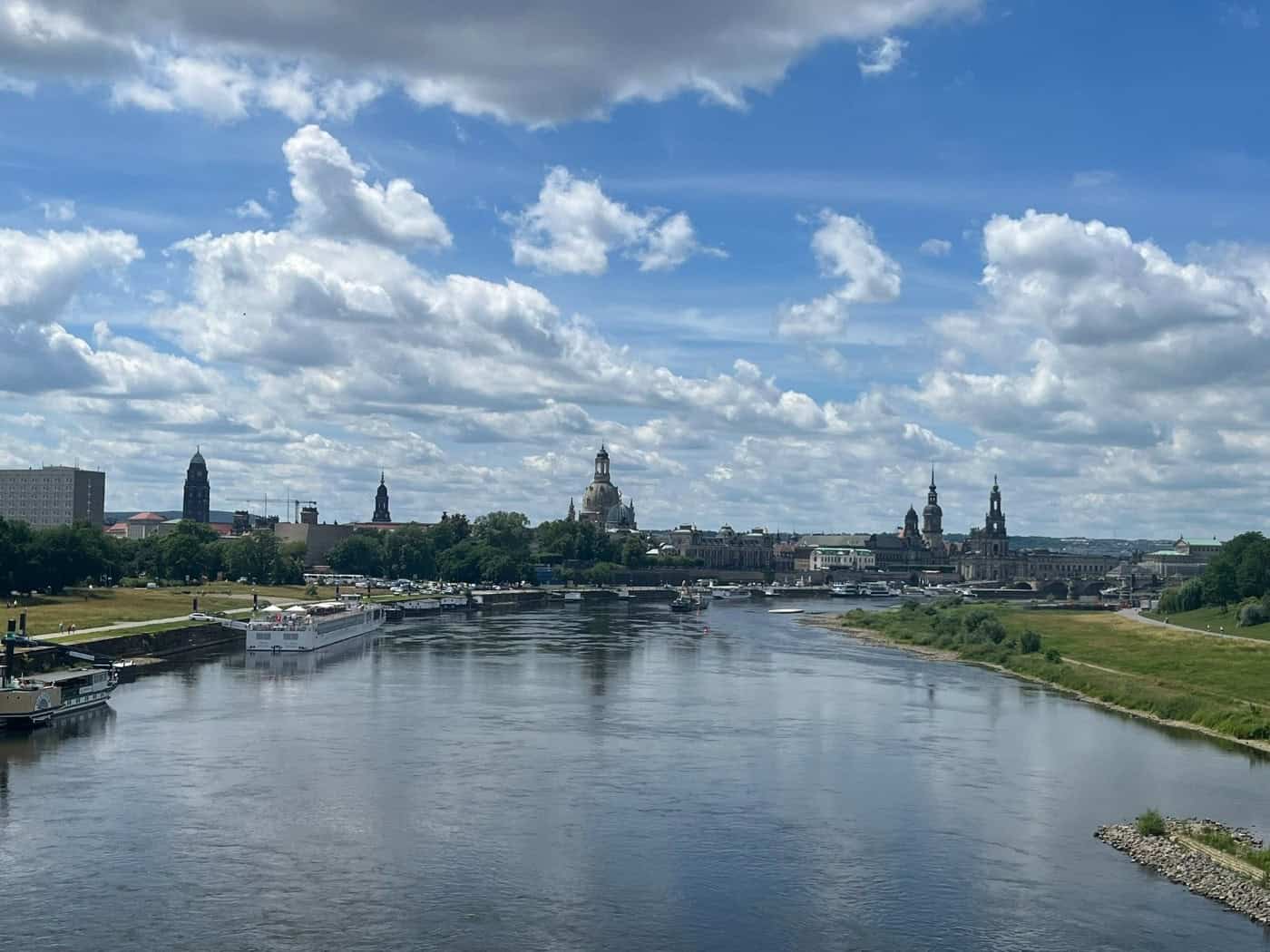 Panorama der Dresdner Altstadt mit Frauenkirche und Hofkirche jenseits der breiten Elbe. Am linken Flussufer liegen weiße Schiffe, rechts erstrecken sich grüne Elbwiesen unter einem leicht bewölkten, blauen Himmel.