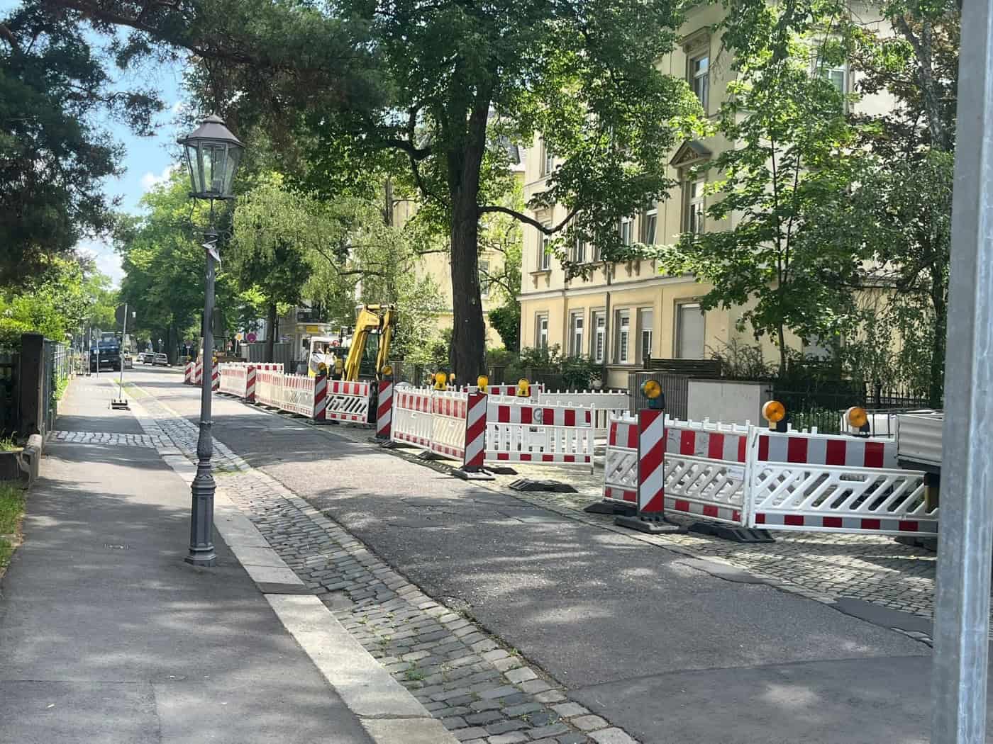 Blick entlang einer Straße mit historischen Villen und großen Bäumen. Die rechte Fahrbahnseite ist mit rot-weißen Gittern abgesperrt, dahinter steht ein gelber Minibagger. Auf dem freien linken Gehweg steht eine klassische Straßenlaterne.
