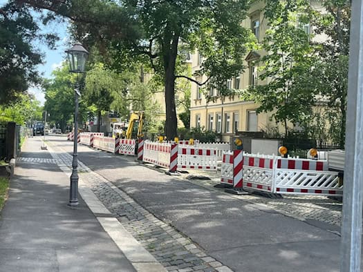 Blick entlang einer Straße mit historischen Villen und großen Bäumen. Die rechte Fahrbahnseite ist mit rot-weißen Gittern abgesperrt, dahinter steht ein gelber Minibagger. Auf dem freien linken Gehweg steht eine klassische Straßenlaterne.