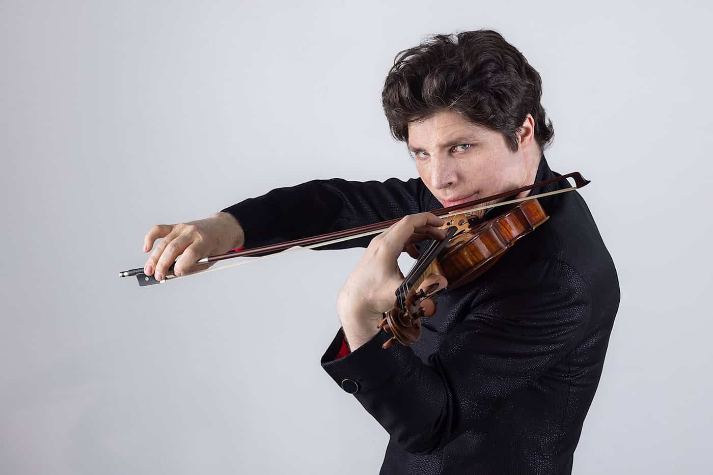 Violin-Solist Augustin Hadelich gastiert in der Semperoper Dresden Eine Porträtaufnahme des Musikers Augustin Hadelich in einem dunklen Anzug. Er hält lächelnd seine Violine und den Bogen in den Händen und blickt freundlich in die Kamera vor einem dunklen, neutralen Hintergrund.