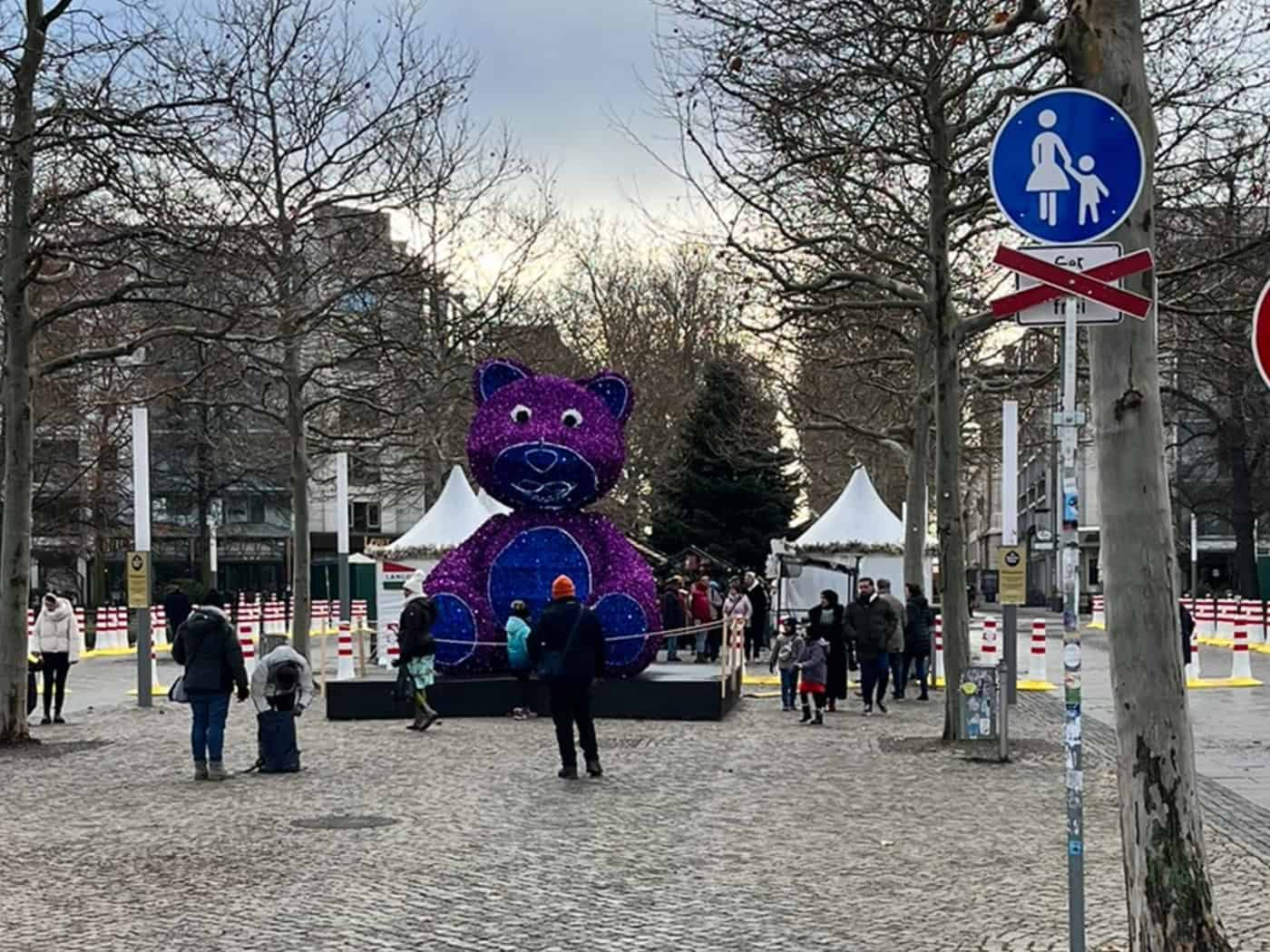 Ein großer, lila-blau leuchtender Teddybär steht auf der gepflasterten Hauptstraße in Dresden. Im Hintergrund des winterlichen Platzes sind kahle Bäume, weiße Marktzelte und ein geschmückter Weihnachtsbaum zu sehen.