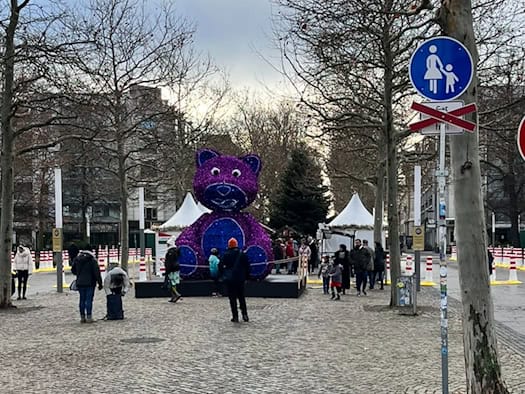 Ein großer, lila-blau leuchtender Teddybär steht auf der gepflasterten Hauptstraße in Dresden. Im Hintergrund des winterlichen Platzes sind kahle Bäume, weiße Marktzelte und ein geschmückter Weihnachtsbaum zu sehen.