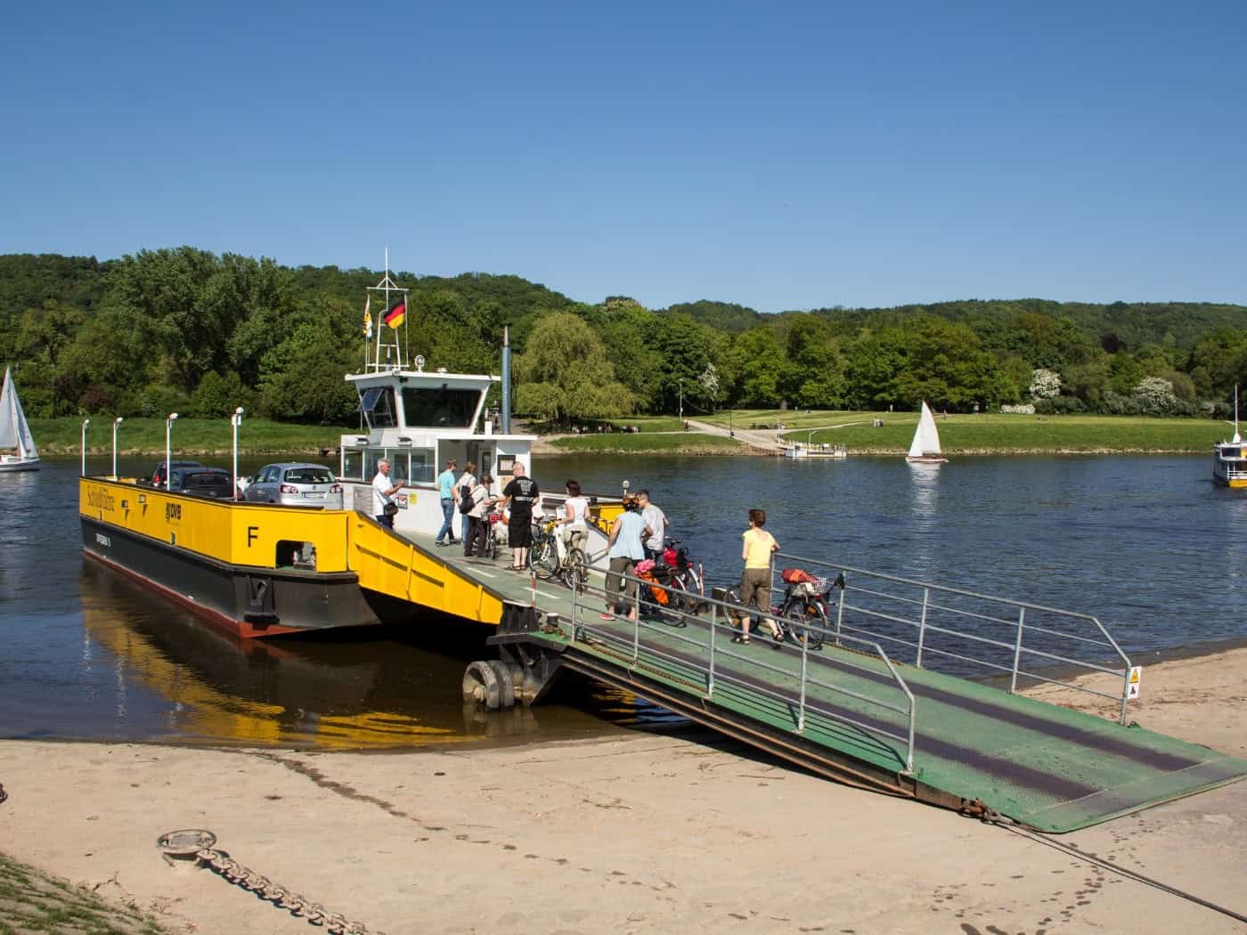 Eine gelb-schwarze Autofähre liegt am sandigen Elbufer. Fahrgäste mit Fahrrädern steigen aus. Auf dem Fluss sind Segelboote vor bewaldeten Hügeln zu sehen.